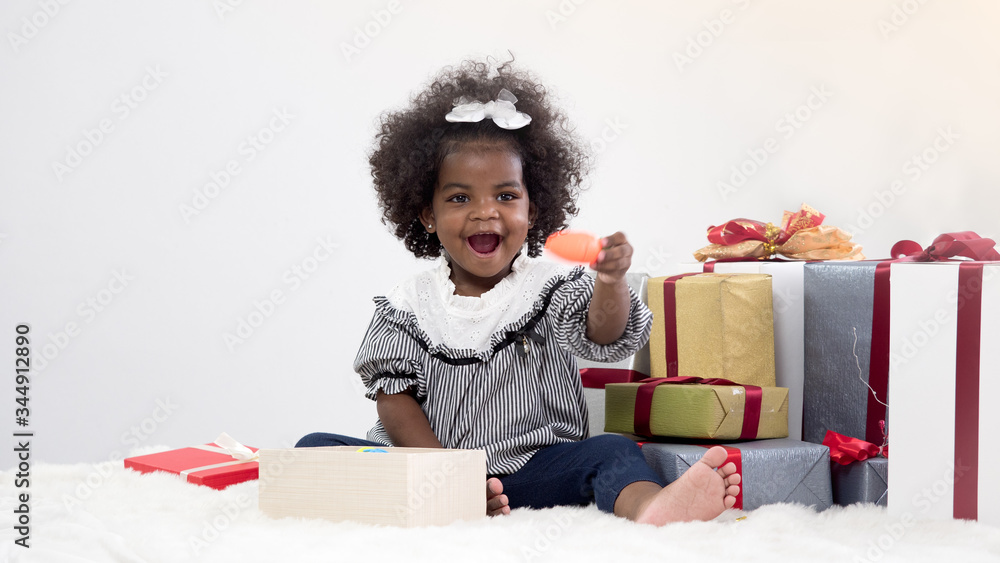 Adorable little African girl sitting next to pile of presents and one opened box. Cute girl holding toy carrot with happiness. Christmas, New Year or Birthday concept. Adorable little African girl sitting next to pile of presents and one opened box. Cute girl holding toy carrot with happiness. Christmas, New Year or Birthday concept.