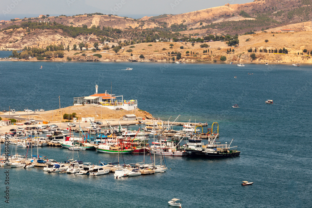 The port of old Foca (in TR: Eski foca limani) with lots of yachts and fishing boats parked. The lighthouse is also visible and several boats are sailing already.