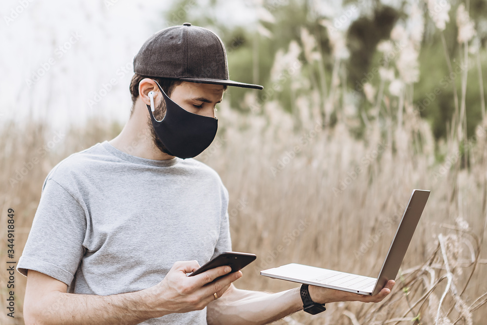 Obraz premium Freelancer during quarantine in the nature. Man in mask is working on laptop. Remote working because of corona virus. Problem of unemployment people. Covid-19 period. Self isolation.