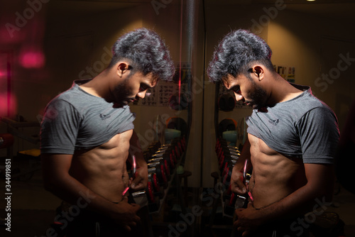 Portrait of black Asian man with beard short hair wear grey shirt show his abs and looking reflection in mirror on fitness background stock photo