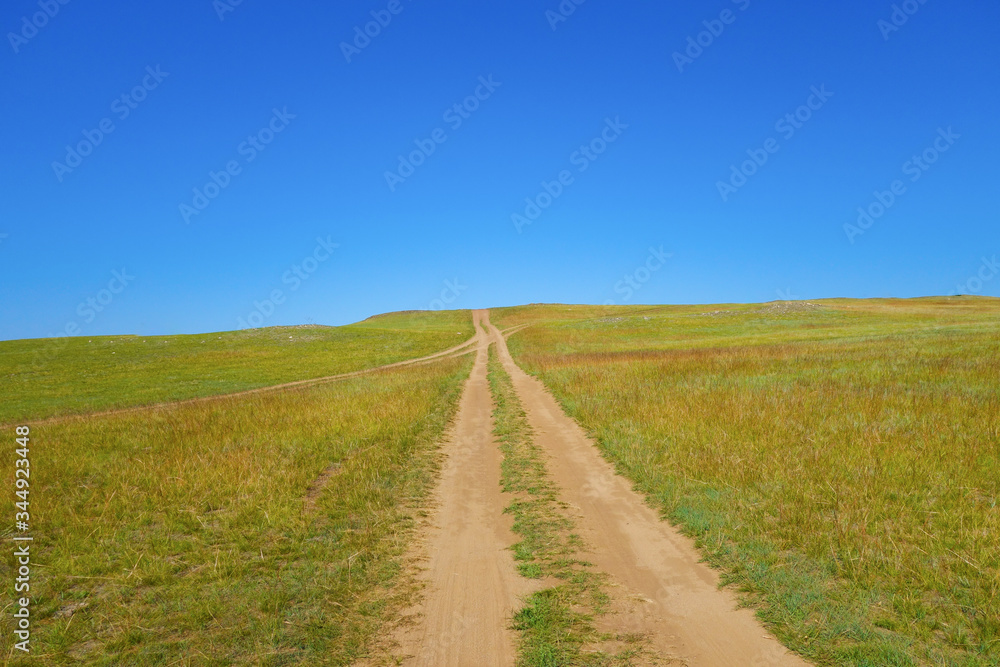 The road to the field goes to the top of the hill. Bright sunny day and cloudless blue sky.