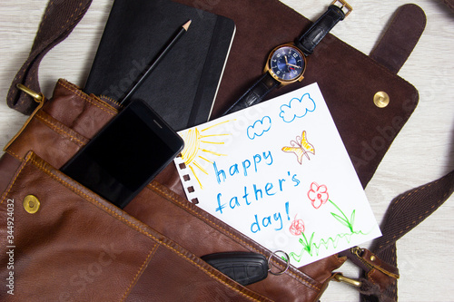 Still life with men's accessories on a wooden table, father's Day concept. Children's drawing b is in the bag next to the clock, Notepad, phone