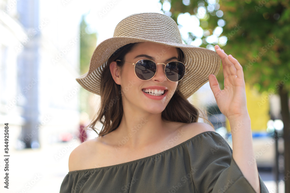 Smiling pretty girl wearing sunglasses and summer hat and looking at camera while walking in city street.