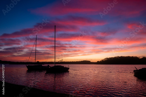 Sunrise in a bay with calm water at high tide over France, sailing ships moored on the shore, visible rocks, islands and trees. Sky in shades of blue, yellow and orange