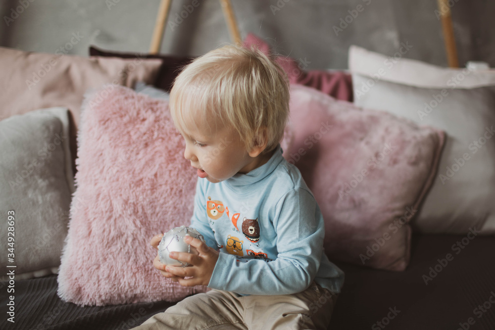 Fototapeta premium A little blond boy with a Christmas ball in his hands is studying the world around him.