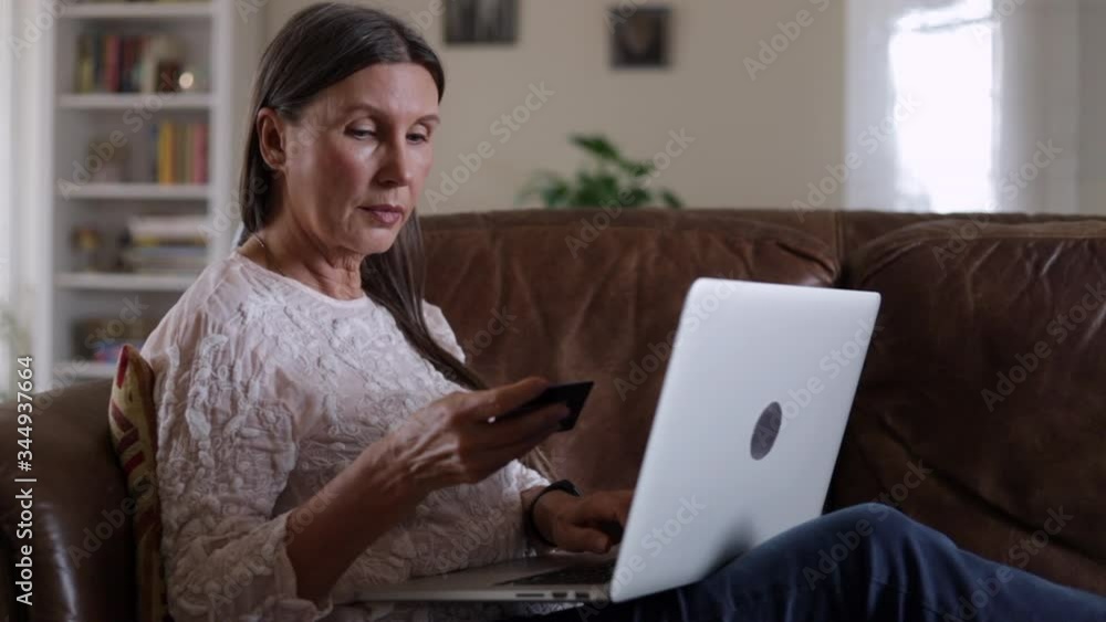Beautiful elderly lady is shopping in online store while sitting on sofa in apartment spbi. Elegant mature woman is making purchases in internet, using credit card and laptop on couch in home interior Beautiful elderly lady is shopping in online store while sitting on sofa in apartment spbi. Elegant mature woman is making purchases in internet, using credit card and laptop on couch in home interior
