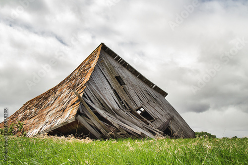 Leinwand Poster An old gray shed built from wood slats collapsed onto green grass in a summer co