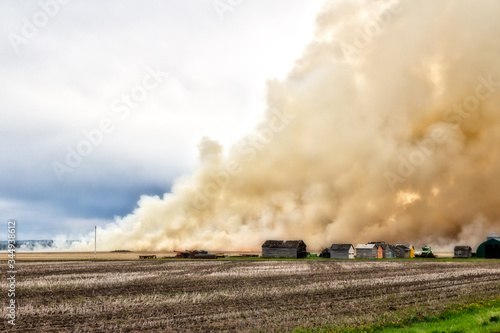 Billowing clouds of smoke from a fire burning a farm field with old sheds nearby in a summer landscape