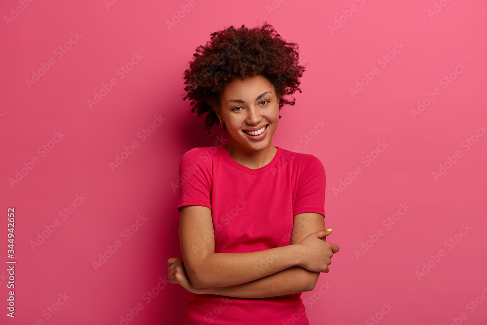 Photo of carefree woman with natural curly hair, keeps hands crossed over body, smiles pleasantly, has casual talk, wears t shirt, enjoys good day, isolated over rosy background. Happy feelings Photo of carefree woman with natural curly hair, keeps hands crossed over body, smiles pleasantly, has casual talk, wears t shirt, enjoys good day, isolated over rosy background. Happy feelings