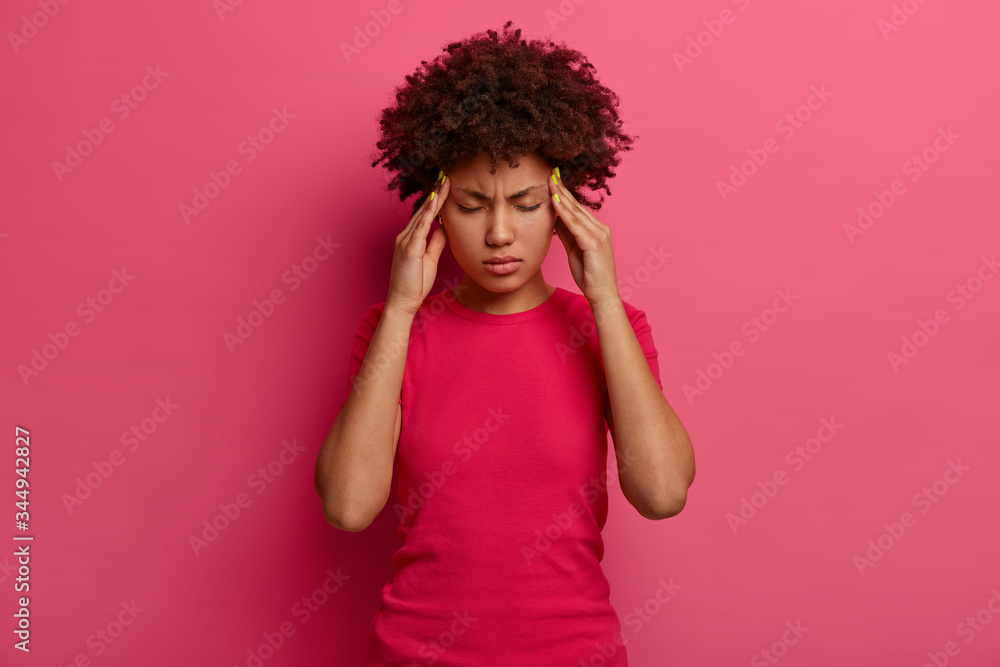 Intense pressed dark skinned millennial woman feels tired and dizzy, keeps hands on temples, suffers from unbearable headache, cannot stands pain, grimaces from migraine, isolated on rosy background Intense pressed dark skinned millennial woman feels tired and dizzy, keeps hands on temples, suffers from unbearable headache, cannot stands pain, grimaces from migraine, isolated on rosy background