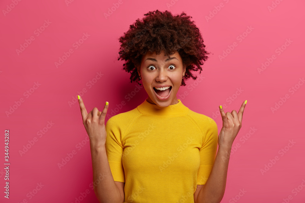 Woman rock n roll star makes heavy metal sign, feels joyful, has bugged eyes and opened mouth, wears yellow t shirt, brings positive vibes, enjoys cool music in club, poses at bright studio. Woman rock n roll star makes heavy metal sign, feels joyful, has bugged eyes and opened mouth, wears yellow t shirt, brings positive vibes, enjoys cool music in club, poses at bright studio.