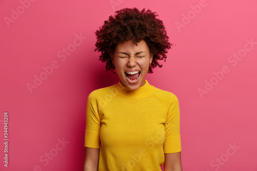 Emotional woman keeps mouth wide opened, yells and screams loudly, cannot control her emotions, being angry with someone, wears yellow t shirt, isolated on pink background, reproaches someone