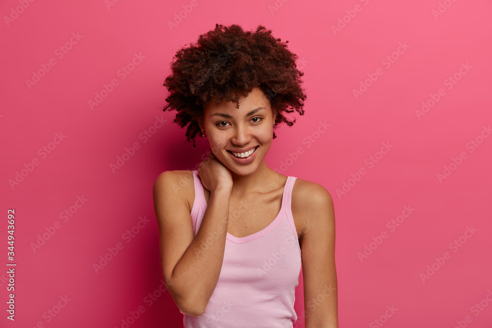 Pleased positive Afro American woman looks directly at camera with charming smile, touches neck gently, feels satisfied, has curly bushy hair, dressed in casual wear, isolated on pink background Pleased positive Afro American woman looks directly at camera with charming smile, touches neck gently, feels satisfied, has curly bushy hair, dressed in casual wear, isolated on pink background