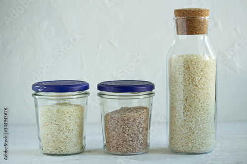 Three types of rice: round, basmati and red in glass jars for storage on a light gray background.