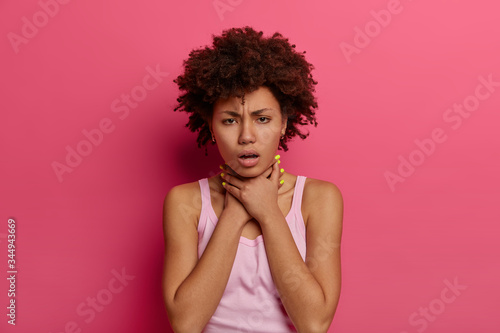 Sick young woman feels unwell, touches neck, suffers from sore throat, has asthmatic fit, cannot breath, has suffocation, poses indoor against rosy background, looks with unhappy expression.
