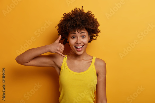 Friendly positive woman shows phone sign, wants to have conversation, keeps contact with close people, speaks to person distantly, dressed in yellow vest, stands indoor, gives her telephone number