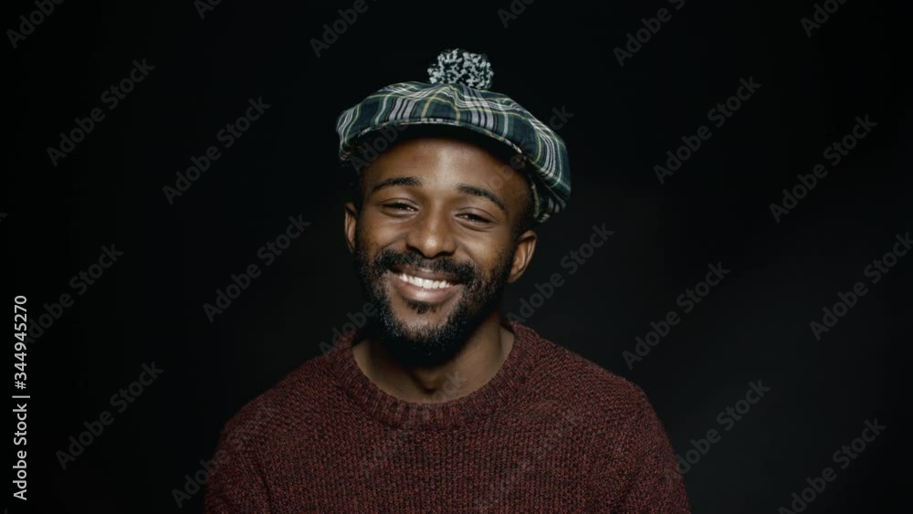 African man in scottish cap smiling against black background. Bearded man in warm clothes looking at camera.
African man in scottish cap smiling against black background. Bearded man in warm clothes looking at camera.