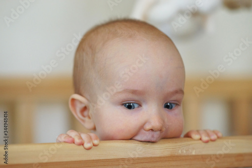 Close portrait of cute newborn child in wooden baby bed at home. Child nibbling wooden beam and looking out of bed