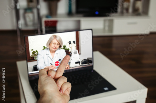 Telemedicine negative concept. Angrey male showing middle finger to a doctor GP on a computer screen, living room home background