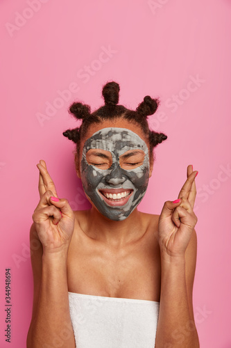 Positive dark skinned woman crosses fingers for good luck, awaits for positive results after spa treatment, applies beauty mask, looks so natural, closes eyes from feeling glad, isolated on pink