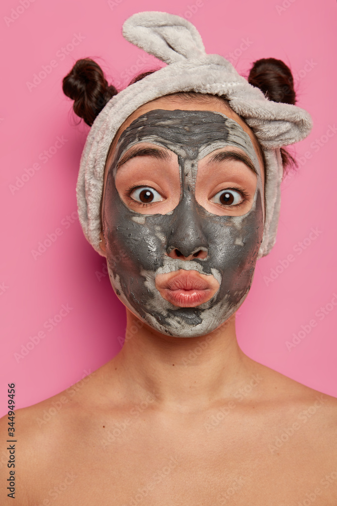 Funny mixed race young woman pouts lips, applies facial clay mask, looks directly at camera, has two combed buns, poses naked indoor, tries new beauty product, glad to have fresh clean skin. Funny mixed race young woman pouts lips, applies facial clay mask, looks directly at camera, has two combed buns, poses naked indoor, tries new beauty product, glad to have fresh clean skin.