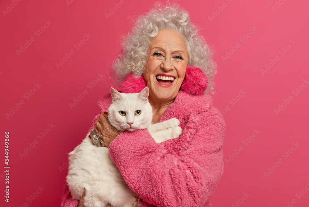 Portrait of aged wrinkled woman enjoys good day with favorite white cat, wears domestic robe, feels not bored on retirement, going to feed pet, loves animals, poses indoor against pink background Portrait of aged wrinkled woman enjoys good day with favorite white cat, wears domestic robe, feels not bored on retirement, going to feed pet, loves animals, poses indoor against pink background