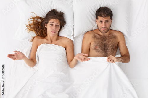Overhead shot of puzzled woman and her husband have sex problems in bed  displeased expressions  lie under white blanket. Man has impotence  erection failure. Daylife family troubles concept