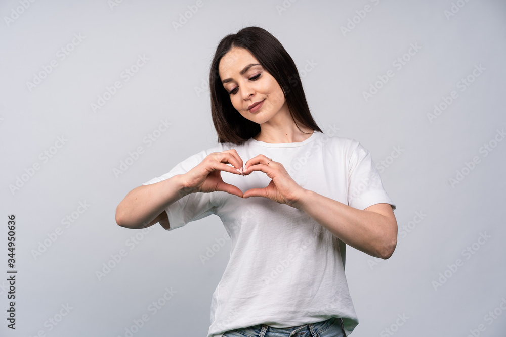 Young beautiful woman shows hands a heart shape, brunette in a white T-shirt on a white background Young beautiful woman shows hands a heart shape, brunette in a white T-shirt on a white background