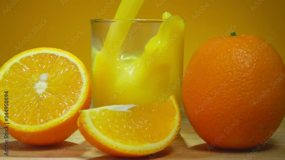 Delicious orange juice is poured in a glass next to large ripe oranges cut into halves on the yellow background in slow motion Delicious orange juice is poured in a glass next to large ripe oranges cut into halves on the yellow background in slow motion