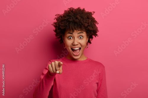Emotional young dark skinned woman looks excited and cheerful, points index finger directly at camera, chooses something or picks someone, wears rosy jumper, poses indoor. People and choice.
