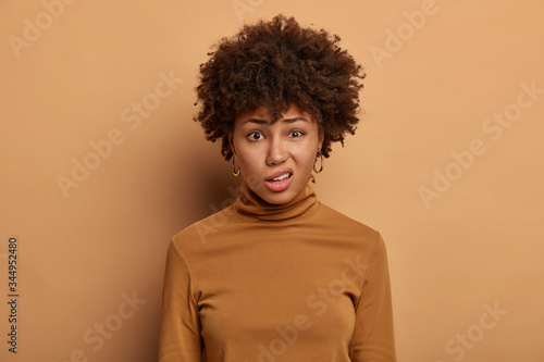 Obraz na plátně Puzzled African American woman has displeased expression, frowns face, looks with disgust at something unpleasant, dressed in casual wear, isolated on brown background