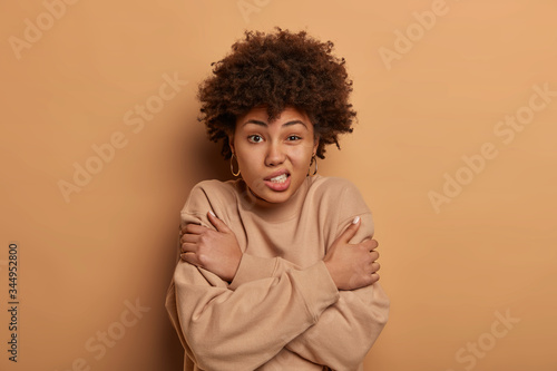 Displeased dark skinned woman hugs herself and trembles, clatters teeth, shakes from cold, asks to lend coat, wears only jumper, smirks face, stands indoor over brown background. It is getting chilly © wayhome.studio 