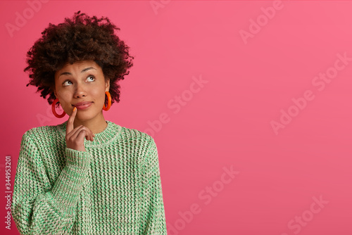 Dreamy dark skinned young woman thinks about career opportunities, stands thoughtful, concentrated upwards, dressed in knitted sweater, isolated on pink background, copy space for your promotion
