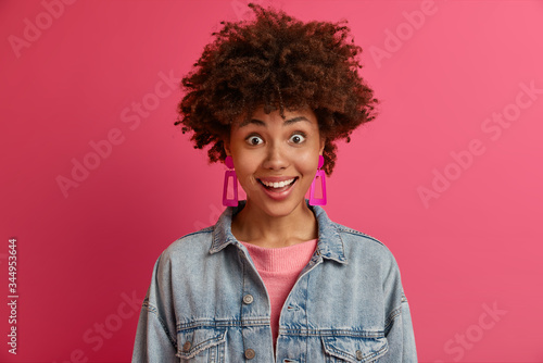 Surprised happy emotional woman with Afro hair looks with smile, cannot believe dreams come true, gets awesome present from someone, dressed in fashionable denim clothes, isolated on pink wall