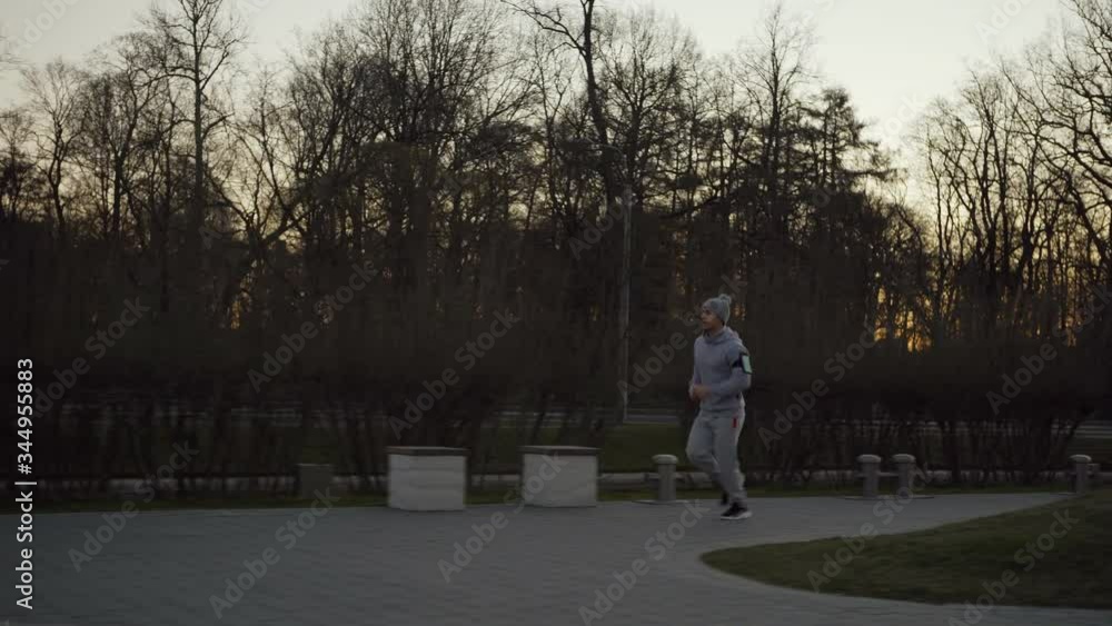 Side view wide tracking shot of young man running down park during jogging practice at sunrise, cell phone on his arm Side view wide tracking shot of young man running down park during jogging practice at sunrise, cell phone on his arm