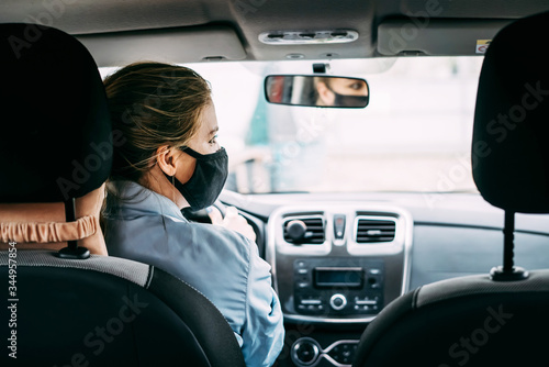 A young taxi driver woman in a black medical mask with blond hair and a turquoise jacket is driving a right-hand drive car. A woman looking at road. Masks protective. Quarantine. Virus. Pandemic.