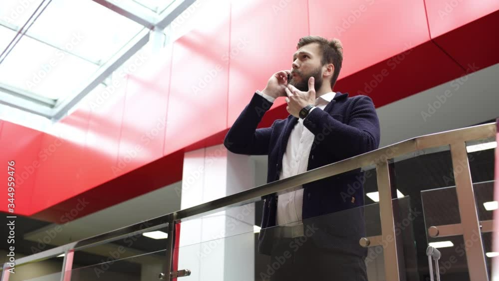 Serious brunette man is talking on the phone while standing near glass fence in big modern building. Young bearded man wearing white shirt and dark jacket takes the call.