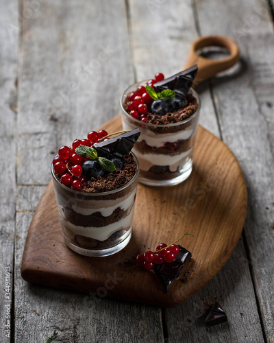 Dessert triffle in transparent glass cups made of chocolate biscuit, chocolate chips, yogurt and berries. Glasses stand on a blackboard, dark photo.