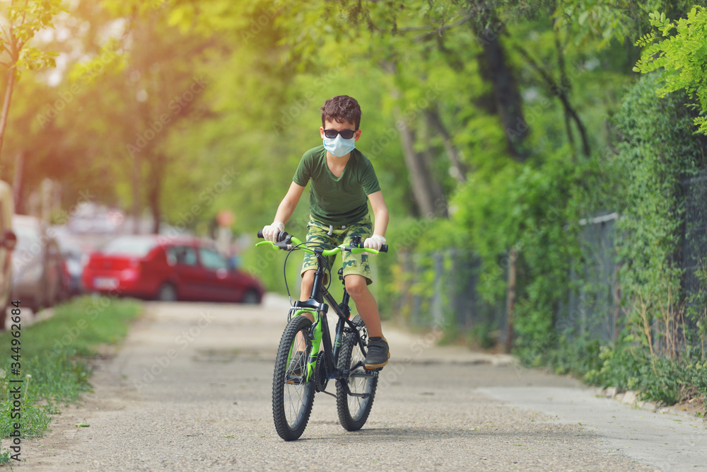 Happy kid having fun near home with a bicycle on beautiful spring day wearing protection mask for coronavirus Covid-19 pandemic virus