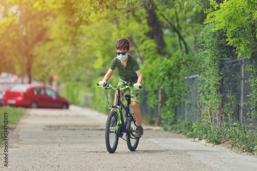 Happy kid having fun near home with a bicycle on beautiful spring day wearing protection mask for coronavirus Covid-19 pandemic virus