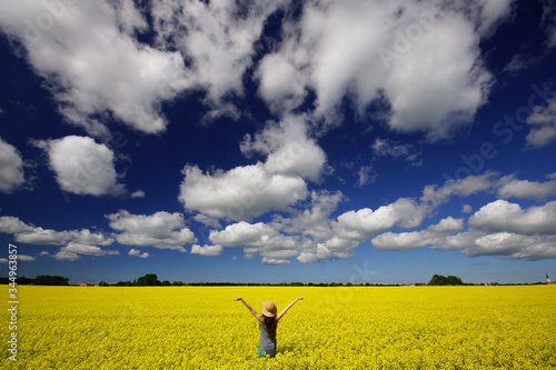 Young woman in straw hat in the middle of bright yellow rapeseed field with hands raised in the air. View from behind