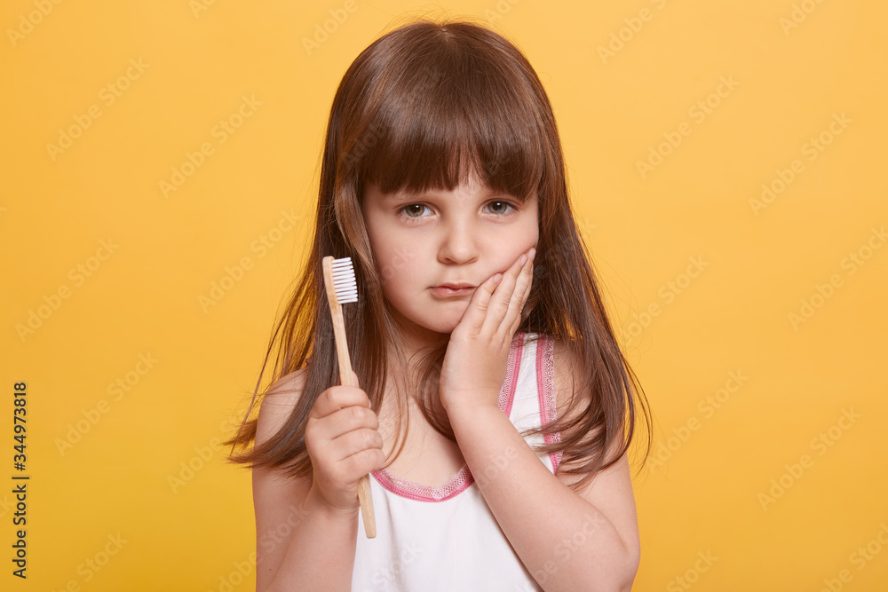 Closeup portrait of serious cute child keeping palm on cheek, having toothache, holding toothbrush in hand, looking at camera, standing isolated over yellow background. Daily dental hygiene concept. Closeup portrait of serious cute child keeping palm on cheek, having toothache, holding toothbrush in hand, looking at camera, standing isolated over yellow background. Daily dental hygiene concept.