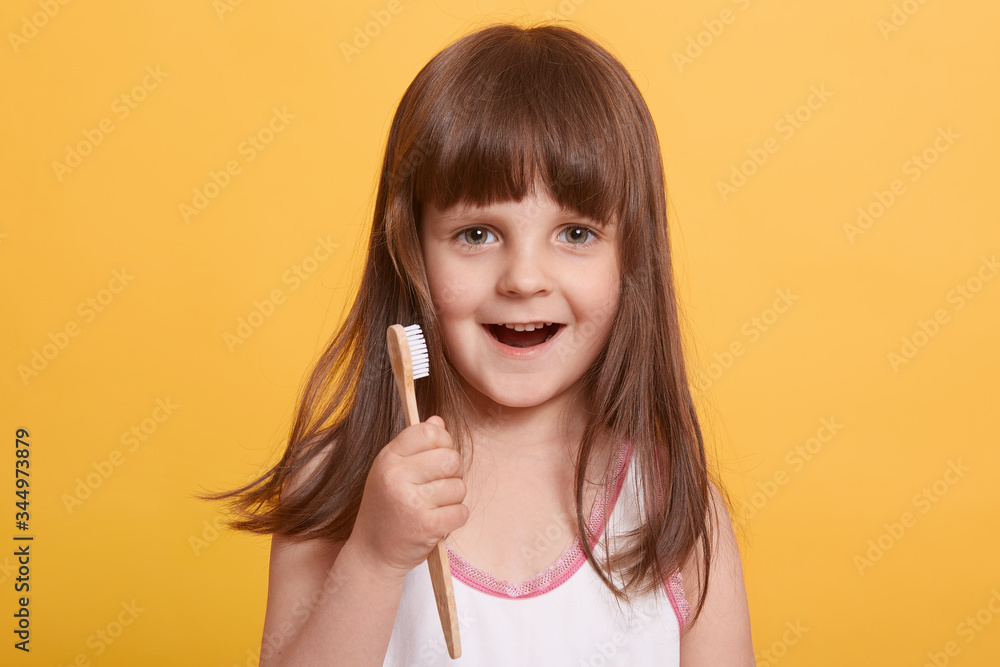 Studio shot of beautiful happy surprised dark haired little girl in bathroom, brushing teeth with excited facial expression, posing isolated over yellow background. Childhood, daily procedures concept Studio shot of beautiful happy surprised dark haired little girl in bathroom, brushing teeth with excited facial expression, posing isolated over yellow background. Childhood, daily procedures concept