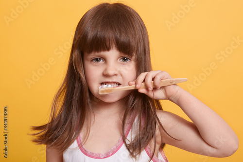 Horizontal shot of charming dark haired female child brushing her teeth, looking directly at camera, having serious emotions, posing isolated over yellow studio background. Daily hygiene concept.