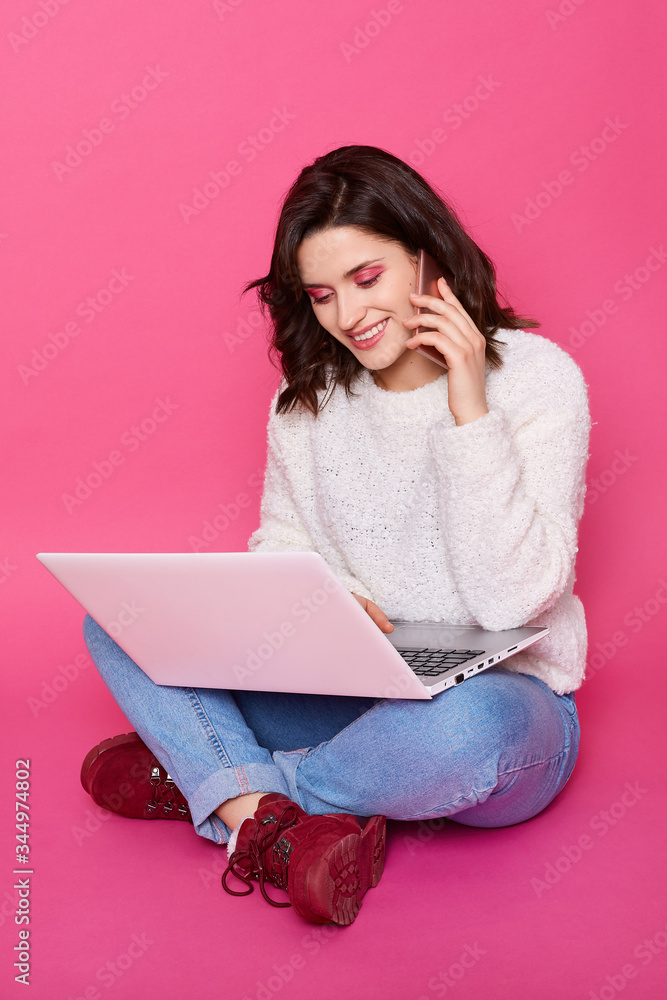 Horizontal shot of smiling woman sitting on floor with lap top and talking to phone, brunette female has pleasant appearance, having positive facial expression, being in good mood, working online. Horizontal shot of smiling woman sitting on floor with lap top and talking to phone, brunette female has pleasant appearance, having positive facial expression, being in good mood, working online.