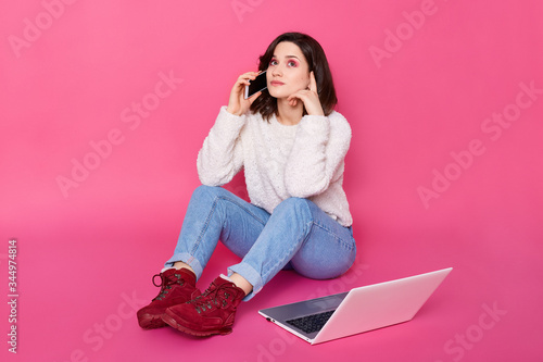 Picture of attractive dark haired girl sitting on floor and talking to friend via smart phone, lady sits near lap top, has pause, work online, girl looking up,has thoughtful facial expression.
