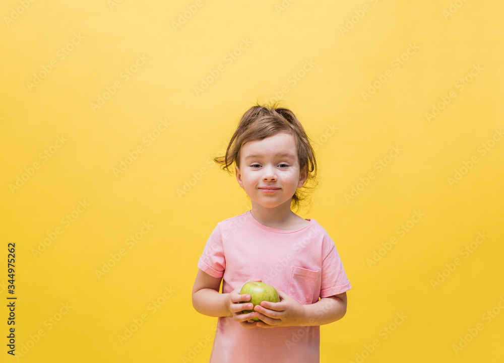 A little girl holding an green Apple on a yellow background. Cute girl with a ponytail in a pink t-shirt. Good nutrition. Copy space A little girl holding an green Apple on a yellow background. Cute girl with a ponytail in a pink t-shirt. Good nutrition. Copy space