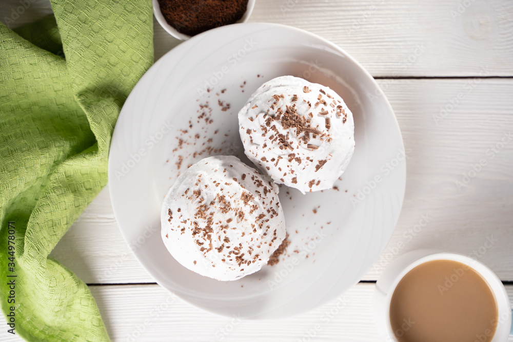 Cupcakes with whipped cream and chocolate chips on a white plate and a Cup of coffee in the background, on a white wooden table. Image for the menu or catalog of confectionery products. Cupcakes with whipped cream and chocolate chips on a white plate and a Cup of coffee in the background, on a white wooden table. Image for the menu or catalog of confectionery products.