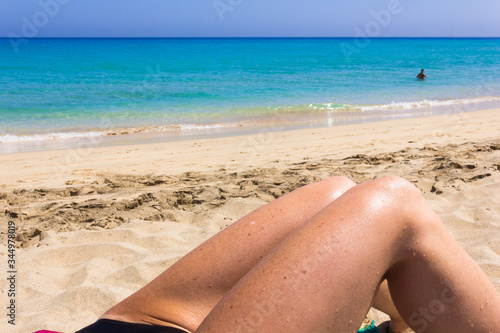 Female legs by white sand beach seashore. Closeup on thigh of woman sunbathing by green and blue water sea. UV protection, permanent hair removal, summer vacation concepts