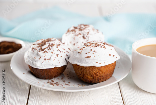 Cupcakes with whipped cream and chocolate chips on a white plate and a Cup of coffee in the background, on a white wooden table. Image for the menu or catalog of confectionery products.